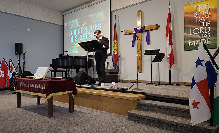 Interior of a church sanctuary with a raised platform, a wooden cross draped with purple cloth, flags displayed along the wall, musical instruments, and a speaker standing at a podium beneath a projected message.