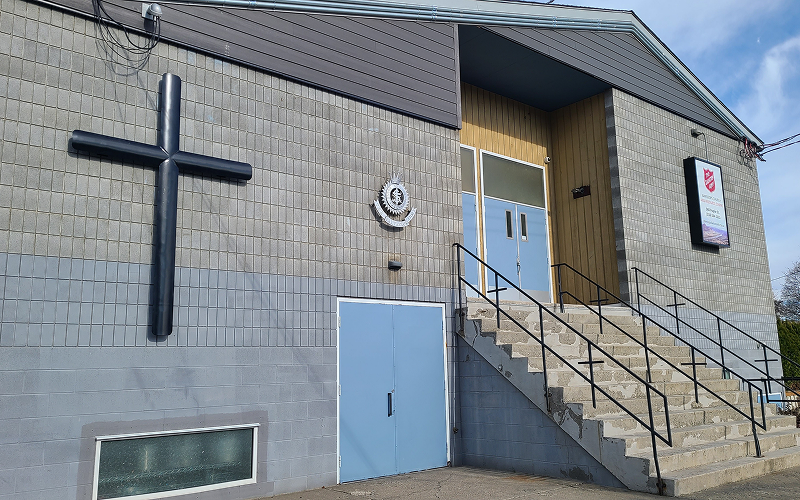Exterior of The Salvation Army Kamloops Church and Resource Centre building, showing the concrete facade with a large cross mounted on the wall and front steps leading to the main entrance.